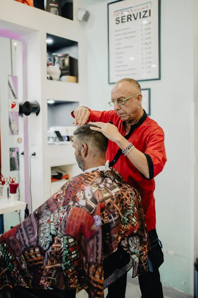Tito taglia i capelli a un cliente, con il listino dei servizi alle spalle
