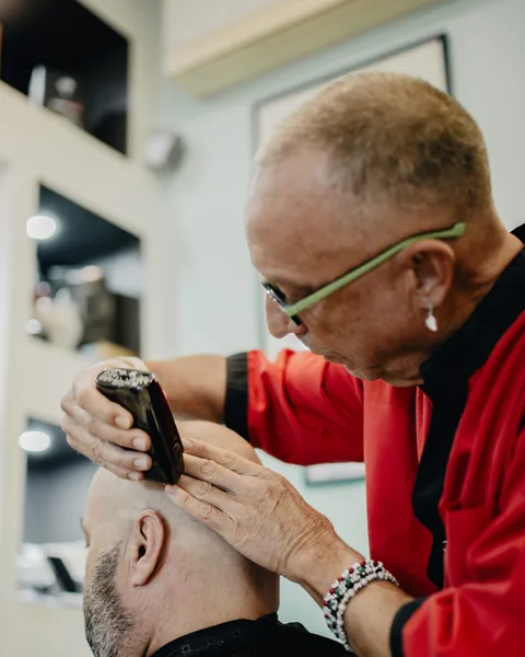 Tito, con gli occhiali verdi, taglia i capelli a un cliente