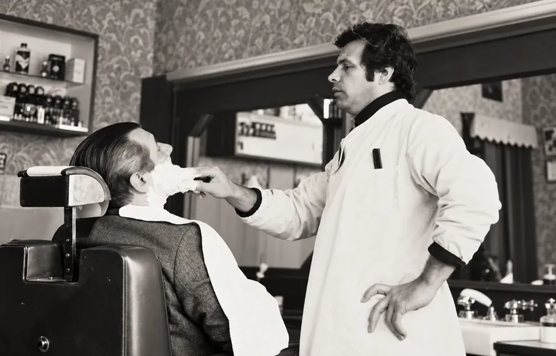 Foto d'epoca in bianco e nero: un barbiere insapona il viso a un cliente